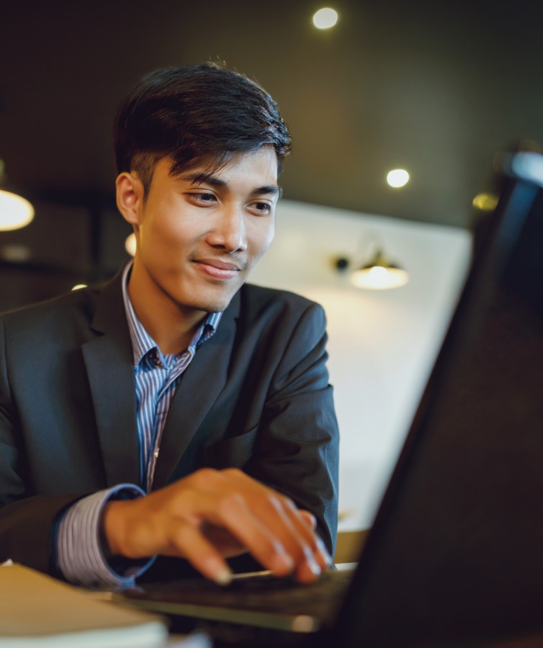 photo of asian man in white and blue striped shirt and a blue suit jacket. He was using a laptop and sitting in an office