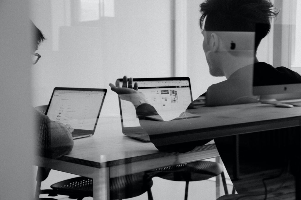 Black and white photos showing two men seated in a conference room looking at laptops.