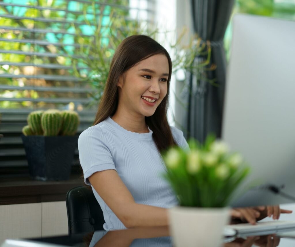 A young woman smiling and working at a computer in a modern office, with potted plants on the desk and sunlight coming through window blinds in the background.