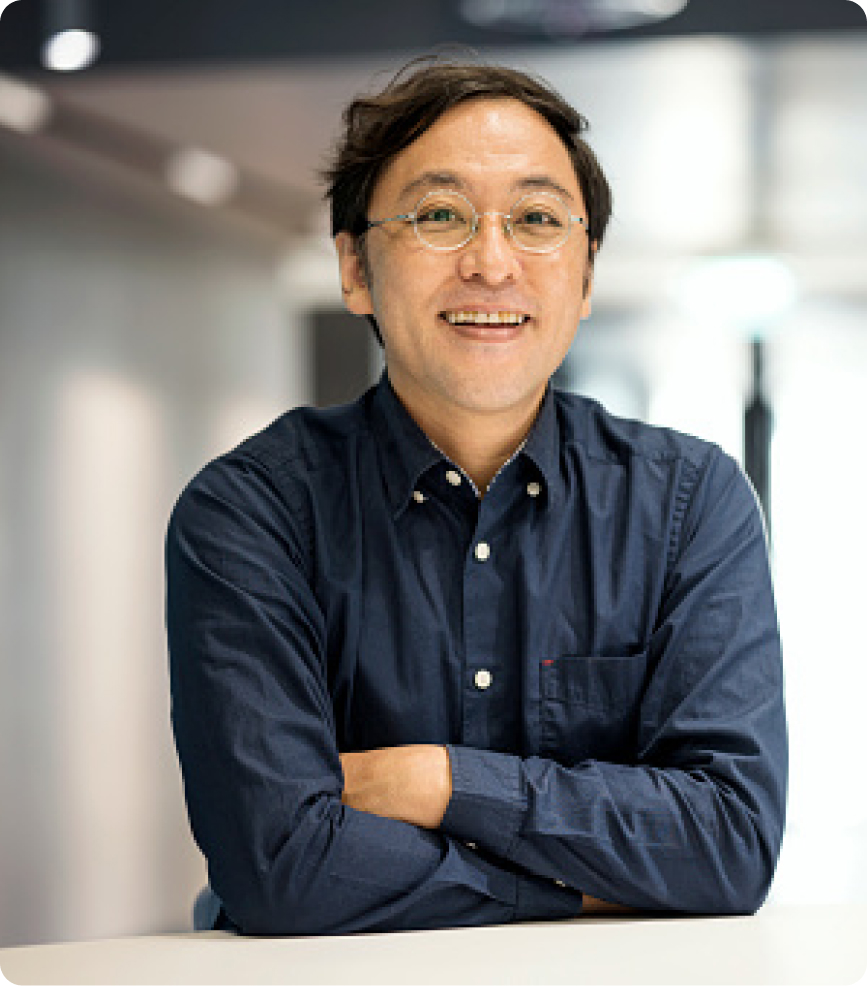 A man with glasses and short dark hair smiles while sitting with arms crossed on a table. He is wearing a navy blue button-up shirt. The background is softly blurred and appears to be an indoor office or workspace.