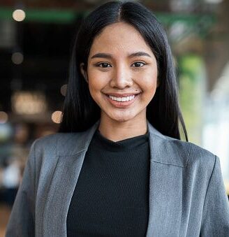 A young woman with long black hair, wearing a gray blazer and black top, smiles confidently while standing indoors in a modern, blurred office or café setting.