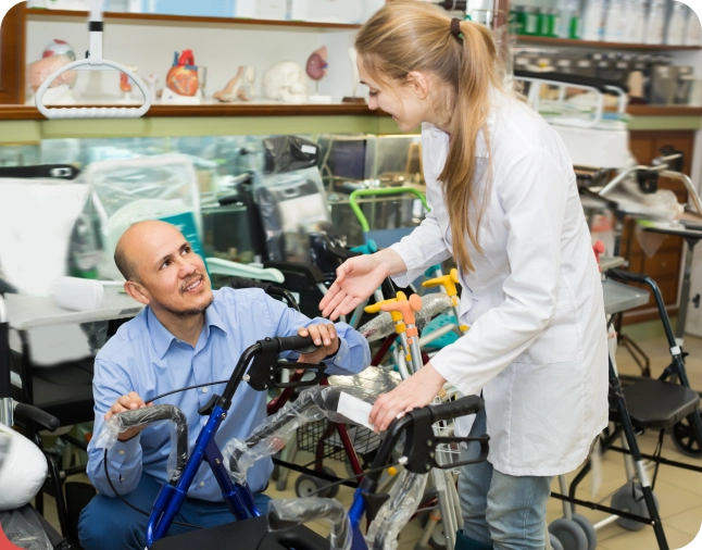 A smiling man tests a blue walker in a medical supply store while a woman in a white coat assists him, gesturing and speaking. Various mobility aids and equipment are displayed around them.