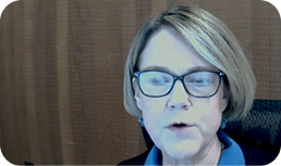 A woman with short light hair and glasses sits in front of a brown background, looking slightly down and speaking. She is wearing a blue top and a black jacket.