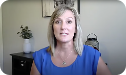 A woman with blonde hair wearing a blue top sits indoors in front of a cabinet with a plant and a decorative item, looking directly at the camera.