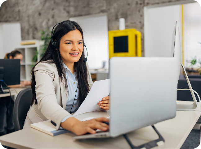 A woman wearing a headset sits at a desk in an office, smiling while looking at her computer screen and holding papers, suggesting she is on a video call or providing customer support.