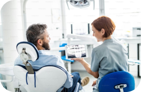 A dentist shows a dental X-ray to a male patient sitting in a dental chair inside a bright, modern dental office.