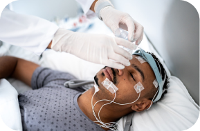 A man lies in a hospital bed with sensors and electrodes attached to his face and head while a healthcare professional adjusts a device on his forehead, likely conducting a sleep study or neurological test.