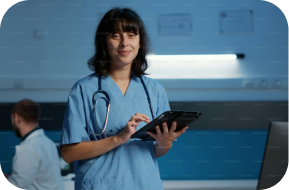 A healthcare professional in blue scrubs with a stethoscope around their neck smiles and uses a tablet in a medical setting, with another person working in the background.