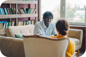 Two people sit facing each other in a cozy room with a bookshelf and large window. One person sits on a sofa, talking and smiling, while the other listens from a chair, seen from behind.