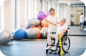 A healthcare worker assists an older adult in a wheelchair with arm exercises using a yellow resistance band in a brightly lit gym with large exercise balls in the background.