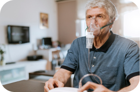 An older man sits indoors using a nebulizer mask for breathing treatment, with medical equipment visible in his hands. The background shows a living room with a television and furniture.