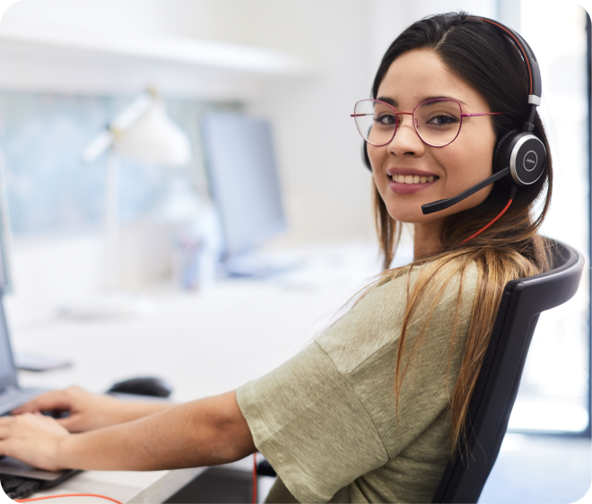 A woman wearing glasses and a headset sits at a desk, smiling at the camera while working on a computer in a bright office environment.