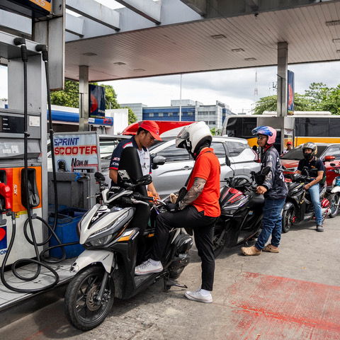 Several people on scooters wait in line at a gas station as an attendant in a red cap—possibly part of the remote staff—assists a rider at the pump. More scooters and cars queue behind, sheltered by the station canopy overhead.