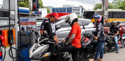Several people on scooters wait in line at a gas station as an attendant in a red cap—possibly part of the remote staff—assists a rider at the pump. More scooters and cars queue behind, sheltered by the station canopy overhead.