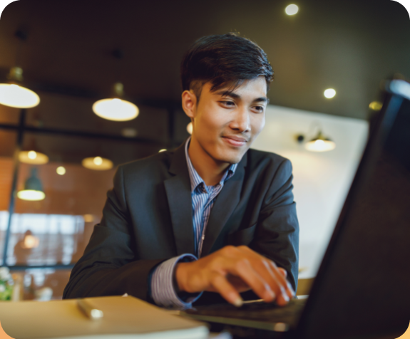 A young man in a suit is smiling while working on a laptop in a modern, warmly lit office or café setting.