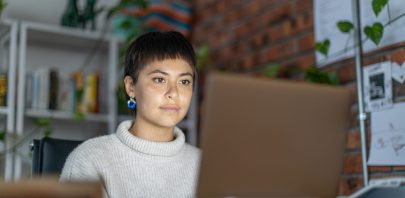 A person with short dark hair and blue earrings sits at a desk, focused on a laptop. They wear a light sweater, and the background shows shelves with books, plants, and a brick wall—ideal for remote staffing security tasks.