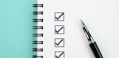 A spiral notebook with four checked boxes on a checklist page and a black pen on the right side, all set against a light blue background—perfect for organizing tasks as you Hire Skilled Back Office Staff in 7 Days.