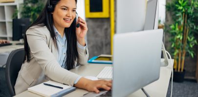 A woman wearing a headset sits at a desk with a notebook, keyboard, and computer. She is smiling and talking, possibly with remote teams or on a customer service call, in a modern office setting with plants in the background.